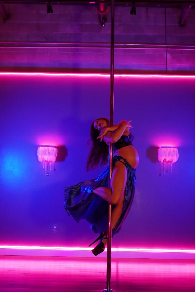 Polina Zueva spinning on the pole with her satin skirt flaring, pregnant, under pink and blue neon.