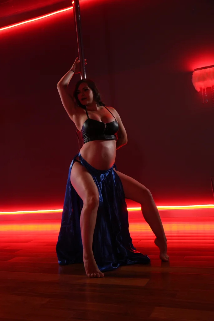 Polina Zueva holding the pole in a lunge under red studio lighting, pregnant, in a blue satin skirt.