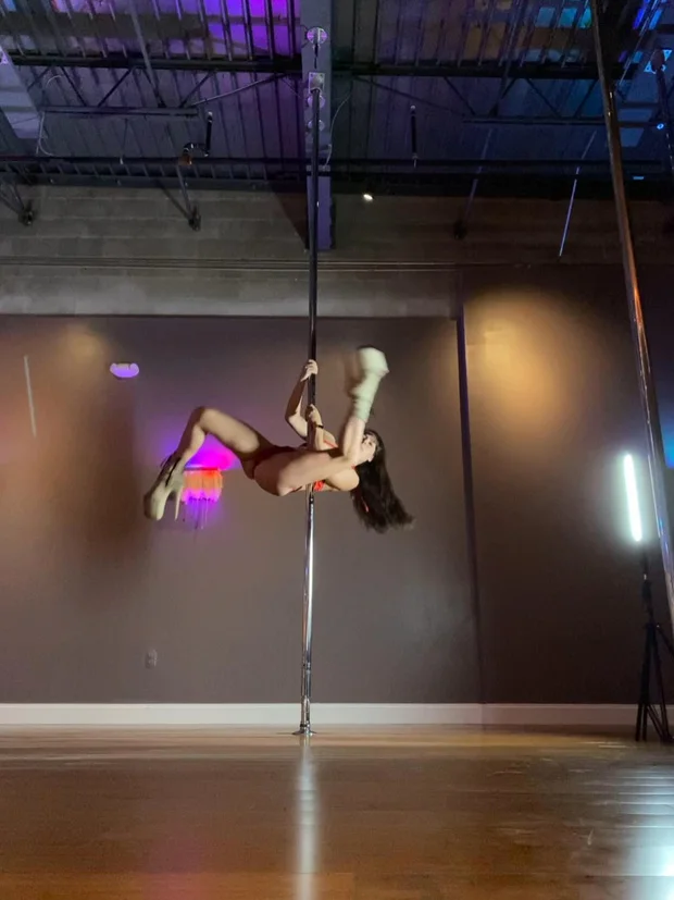 Polina Zueva horizontal on the pole in an orange two-piece, holding the handstand helicopter.