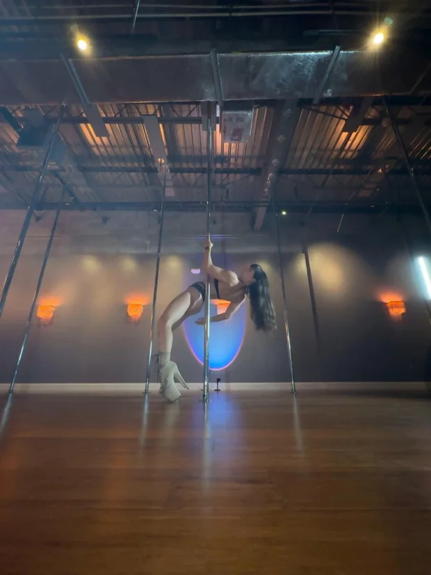 Polina Zueva inverted against the pole in white boots, moving from handstand to forearm stand.