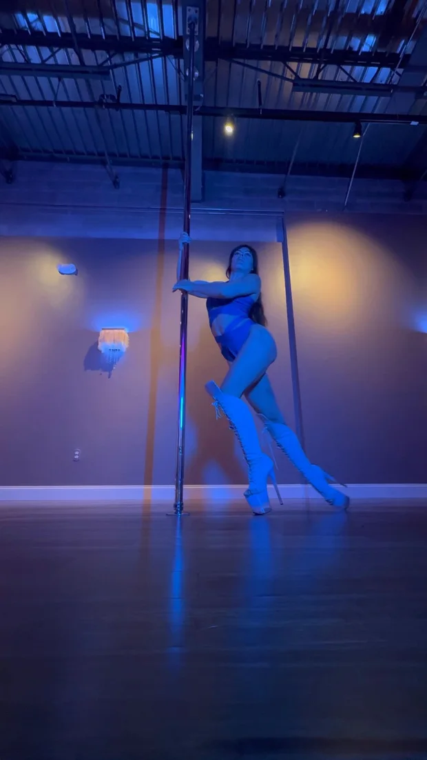 Polina Zueva standing at the pole in blue light and white boots, from Exotic Choreography 1.