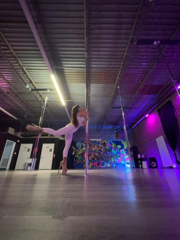 Polina Zueva in a white catsuit with one leg extended along the pole, from Exotic Choreography 4.