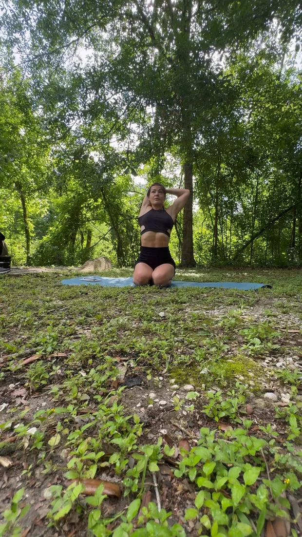 Polina Zueva kneeling on a mat outdoors in a shoulder mobility stretch, trees behind her.
