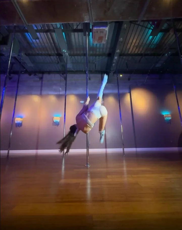 Polina Zueva inverted mid-movement on the pole in white boots, from the forward flip-style jump lesson.